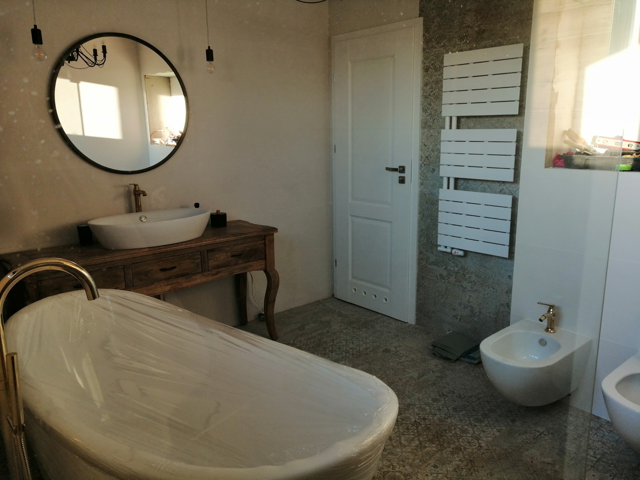 Nowoczesna łazienka w trakcie wykańczania: wolnostojąca wanna zabezpieczona folią, umywalka nablatowa na drewnianej konsoli, bidet i grzejnik drabinkowy na tle wzorzystej ściany.