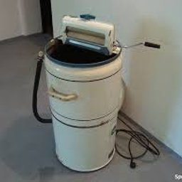 serwisagd - Staromodna pralka wirnikowa w beżowym kolorze z wyżymaczką, stojąca w rogu pomieszczenia z szarą podłogą i białymi ścianami, widoczne przewody i wąż odpływowy.