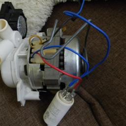 serwisagd - Pompa z silnikiem i kondensatorem zdemontowana z urządzenia AGD, widoczne przewody elektryczne i elementy mocujące.