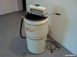 Staromodna pralka wirnikowa w beżowym kolorze z wyżymaczką, stojąca w rogu pomieszczenia z szarą podłogą i białymi ścianami, widoczne przewody i wąż odpływowy.