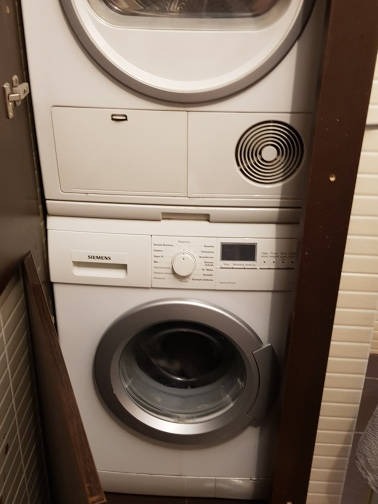 Biała pralka Siemens, ustawiona pod białą suszarką w łazience z białymi płytkami, widok z boku, z fragmentem brązowej płyty po prawej stronie.