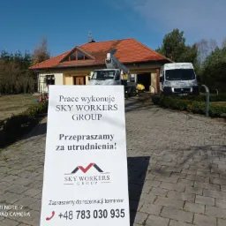 Dom jednorodzinny z częściowo wyczyszczonym dachem z czerwonej dachówki, widoczny z podjazdu wyłożonego kostką brukową, na którym stoi samochód z podnośnikiem oraz tablica informacyjna firmy Sky...