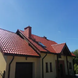 Dom jednorodzinny z nową, czerwoną dach&oacute;wką ceramiczną, widoczny komin z cegły, rynny w kolorze grafitowym, błękitne niebo.