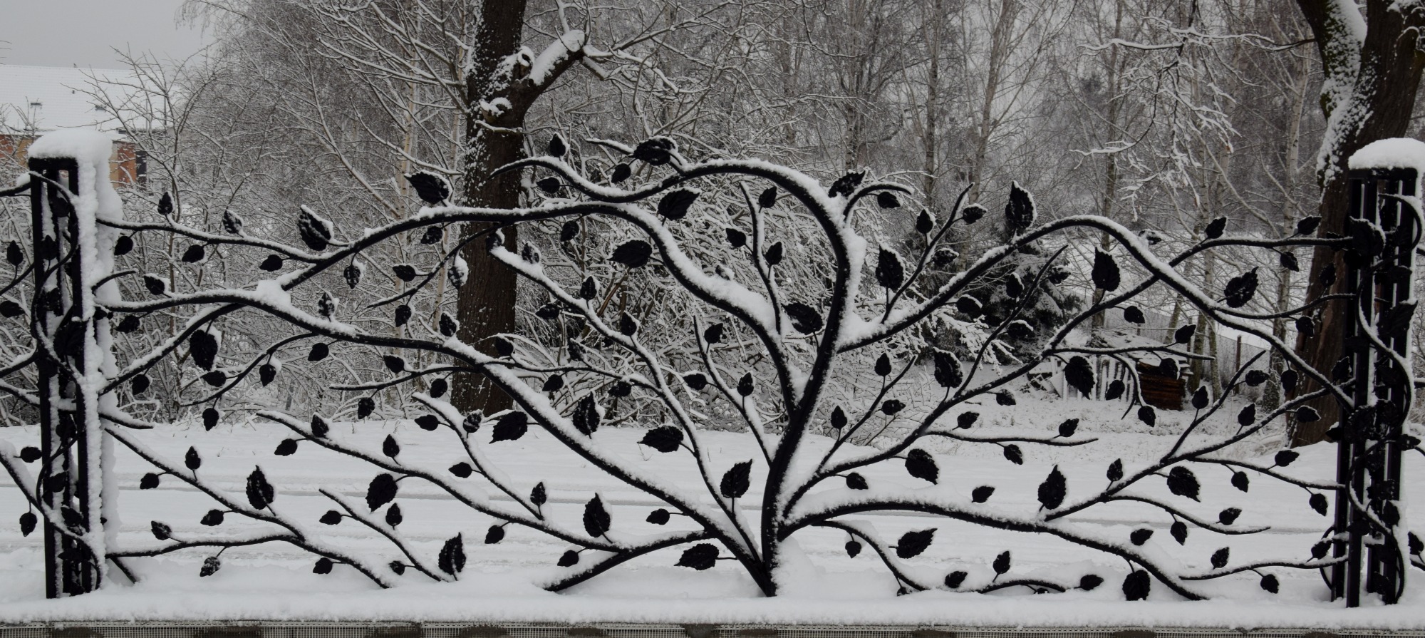 Kute ogrodzenie panelowe z motywem drzewa i liści pokryte śniegiem, na tle zimowego krajobrazu.