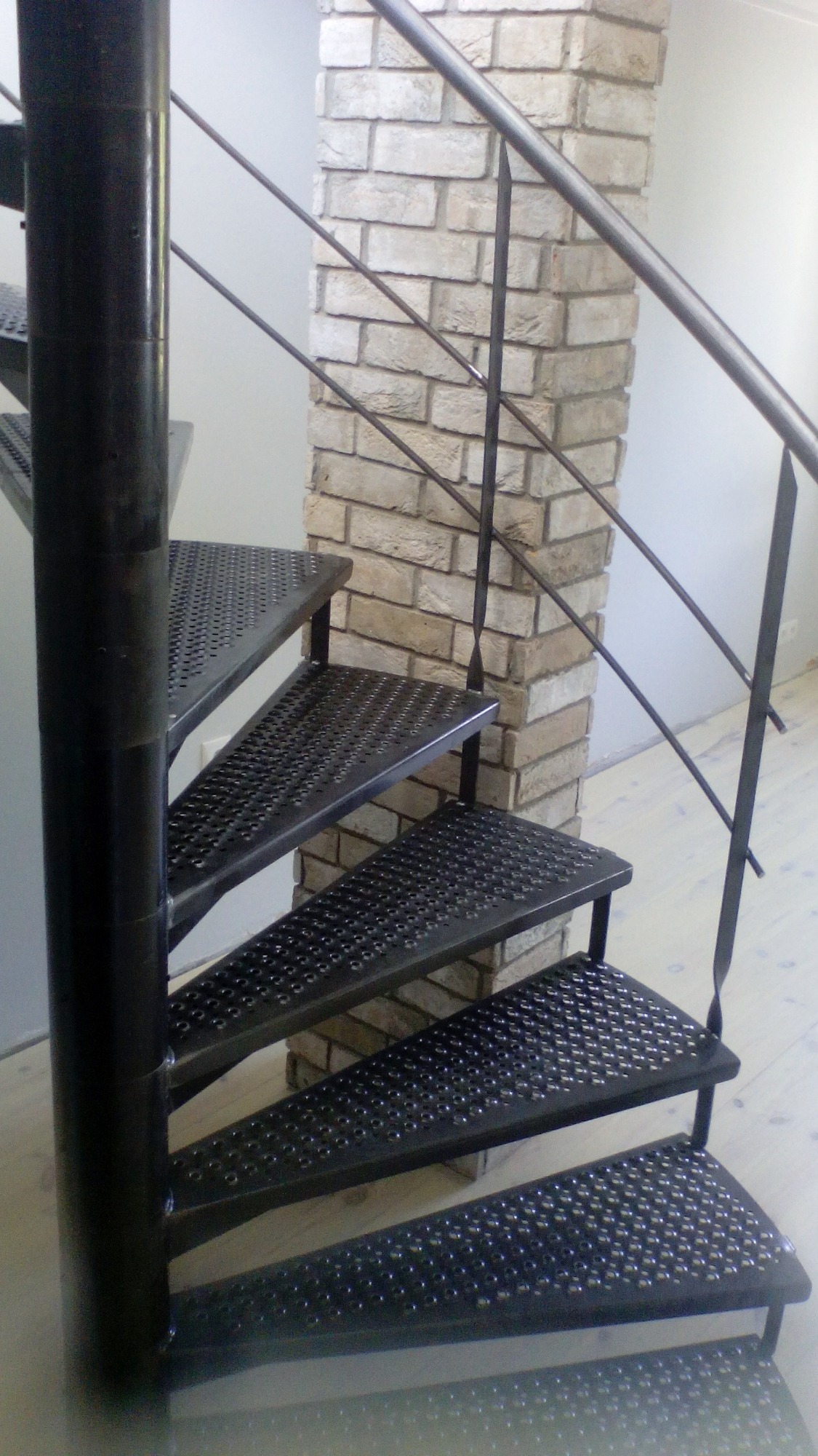 Spiralne schody metalowe z perforowanymi stopniami i czarną balustradą, ustawione na tle ceglanej kolumny w jasnym wnętrzu.