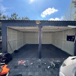 OPIANKOWANI - ocieplenia i izolacje natryskowe pianką PUR - Ocieplony garaż blaszany z otwartymi wrotami, widoczne ślady pianki na podłożu. W tle zieleń drzew i błękitne niebo. Ujęcie z zewnątrz, z bliska.