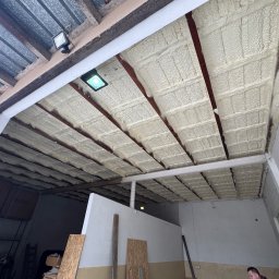 OPIANKOWANI - ocieplenia i izolacje natryskowe pianką PUR - Ocieplenie poddasza pianką PUR, widoczne równomierne pokrycie między krokwiami. Oświetlenie LED w tle. Częściowo widoczne płyty OSB i fragment sylwetki osoby.