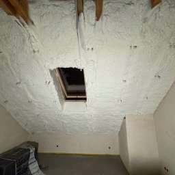 OPIANKOWANI - ocieplenia i izolacje natryskowe pianką PUR - Poddasze po natrysku izolacji pianką PUR, widoczne drewniane elementy konstrukcyjne i okno dachowe. Biała, porowata struktura pianki pokrywa skosy i sufit.