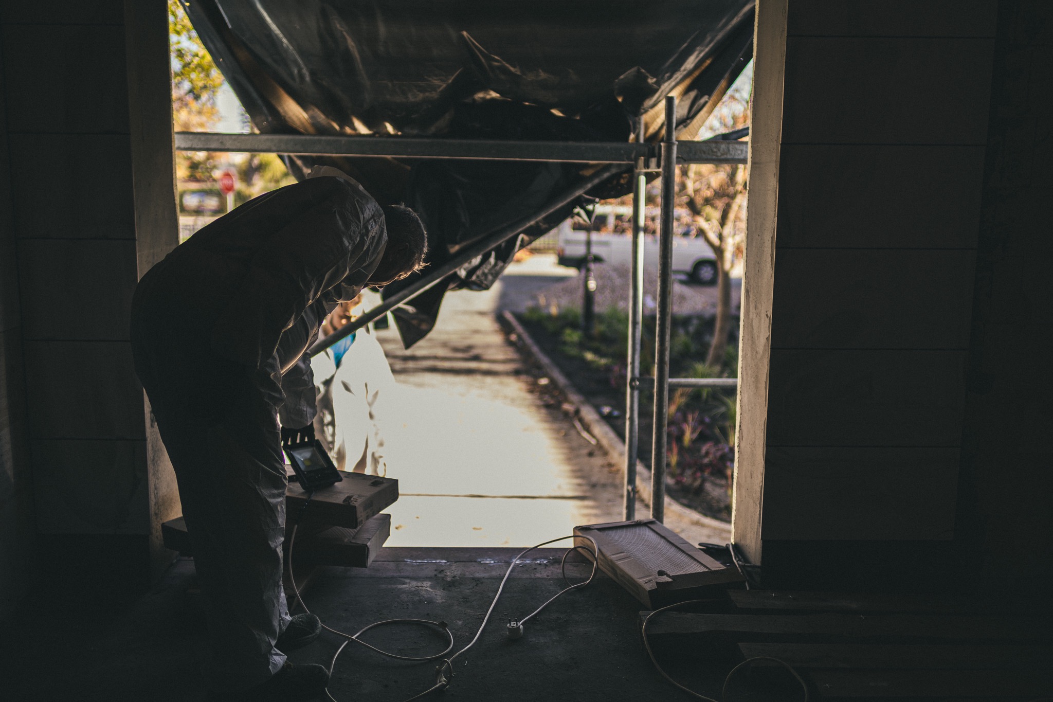 Pracownik w kombinezonie ochronnym pochylony nad sprzętem kontrolnym na budowie, widoczne rusztowanie i okrycie ochronne.