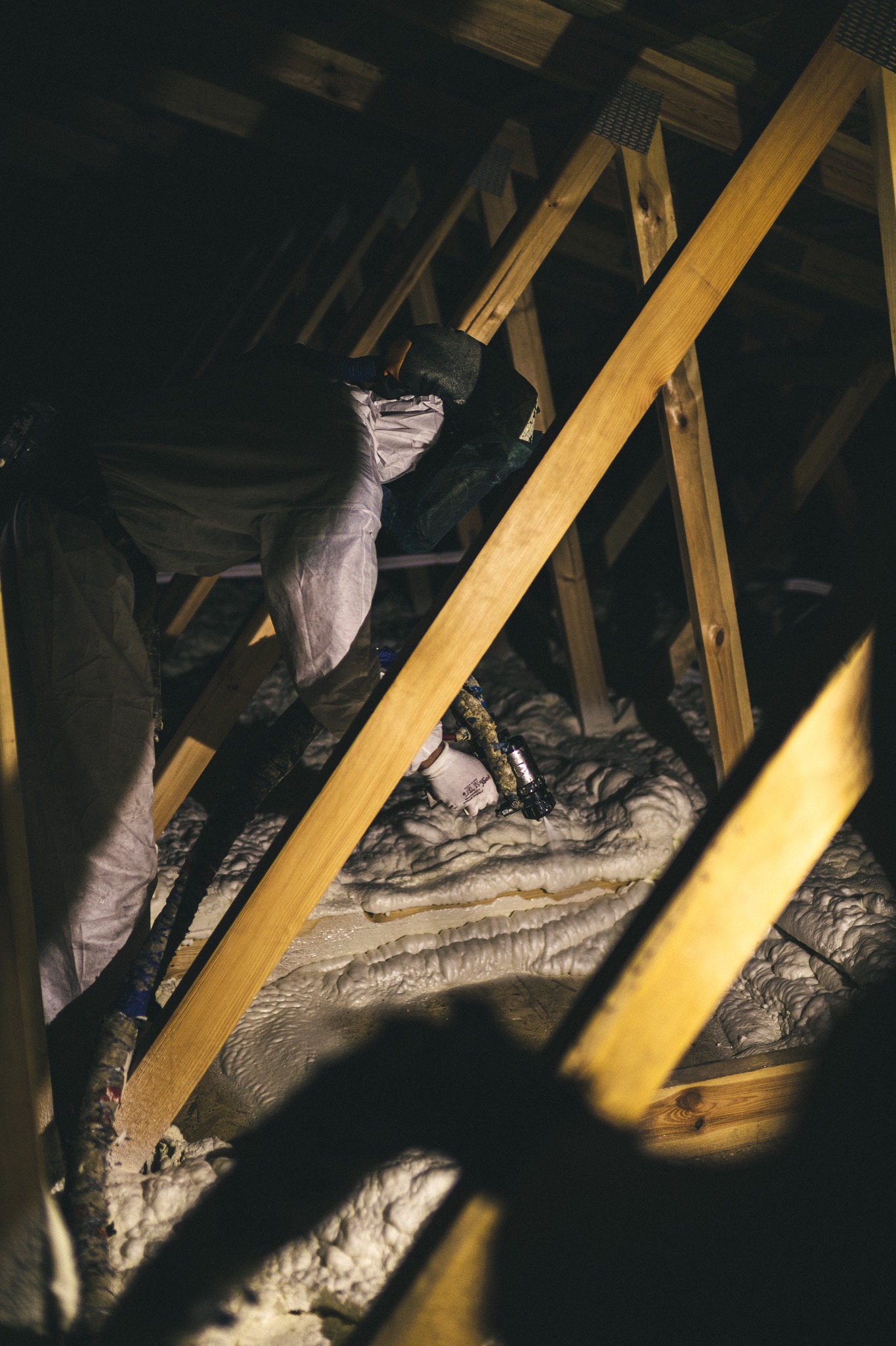 Pracownik w kombinezonie ochronnym aplikuje izolację natryskową w przestrzeni poddasza, widoczne drewniane krokwie i częściowo pokryta powierzchnia.