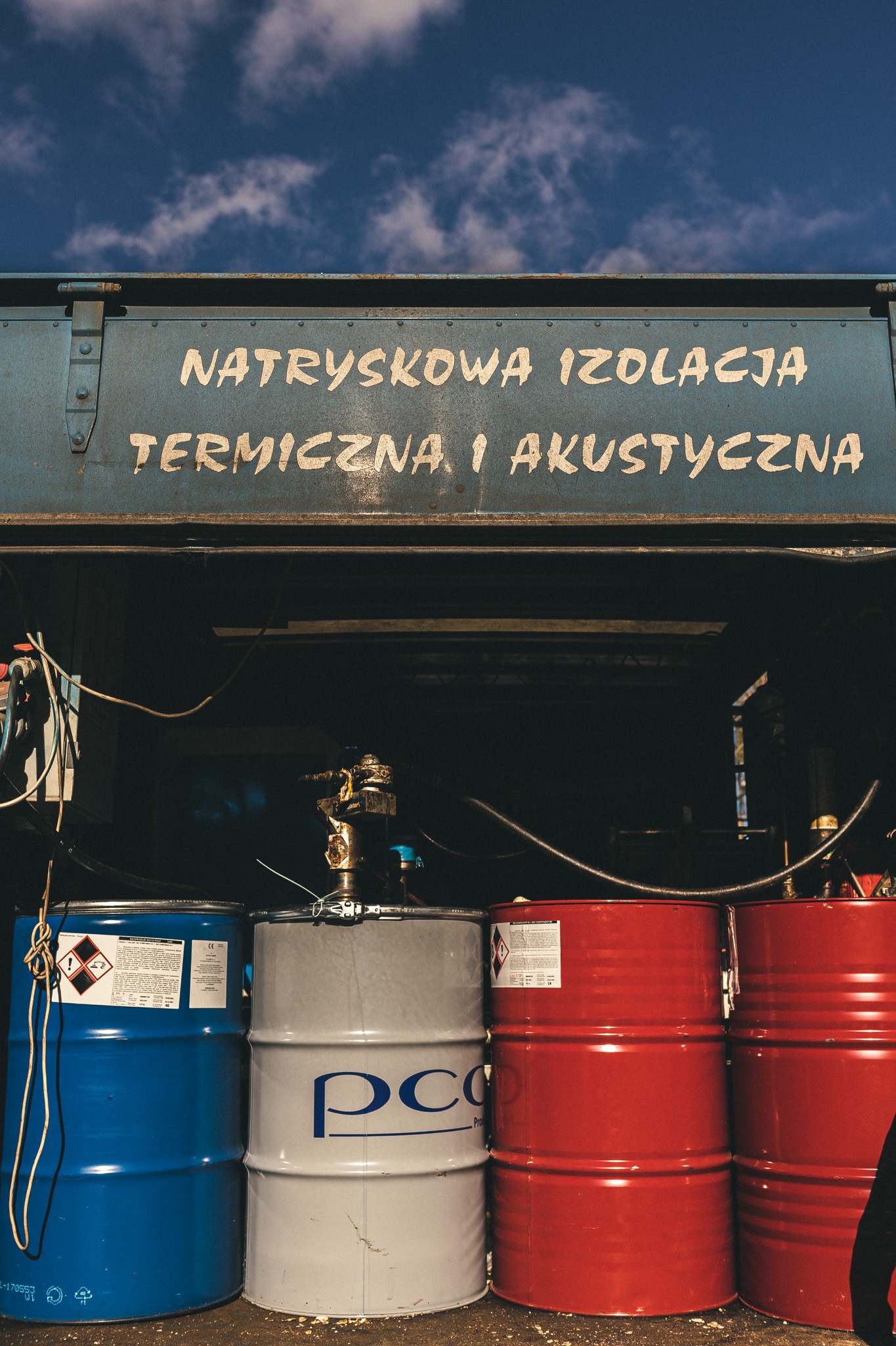 Beczki z materiałami do natryskowej izolacji termicznej i akustycznej, w kolorach niebieskim, szarym i czerwonym, ustawione przed maszyną natryskową z napisem.