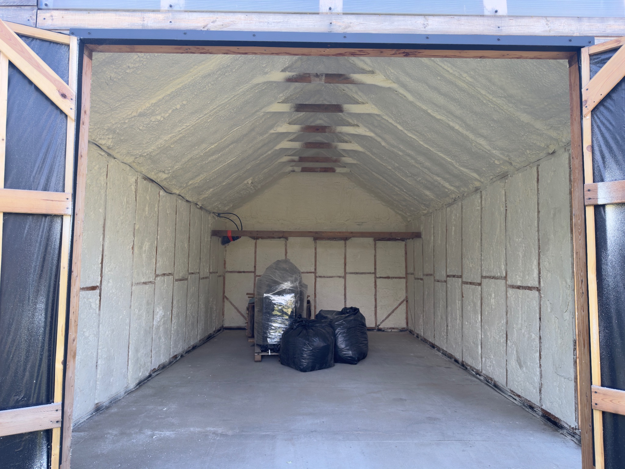 Ocieplony pianką PUR garaż z otwartymi drzwiami, widoczne drewniane elementy konstrukcyjne i czarne worki na podłodze. Izolacja natryskowa ścian i sufitu.