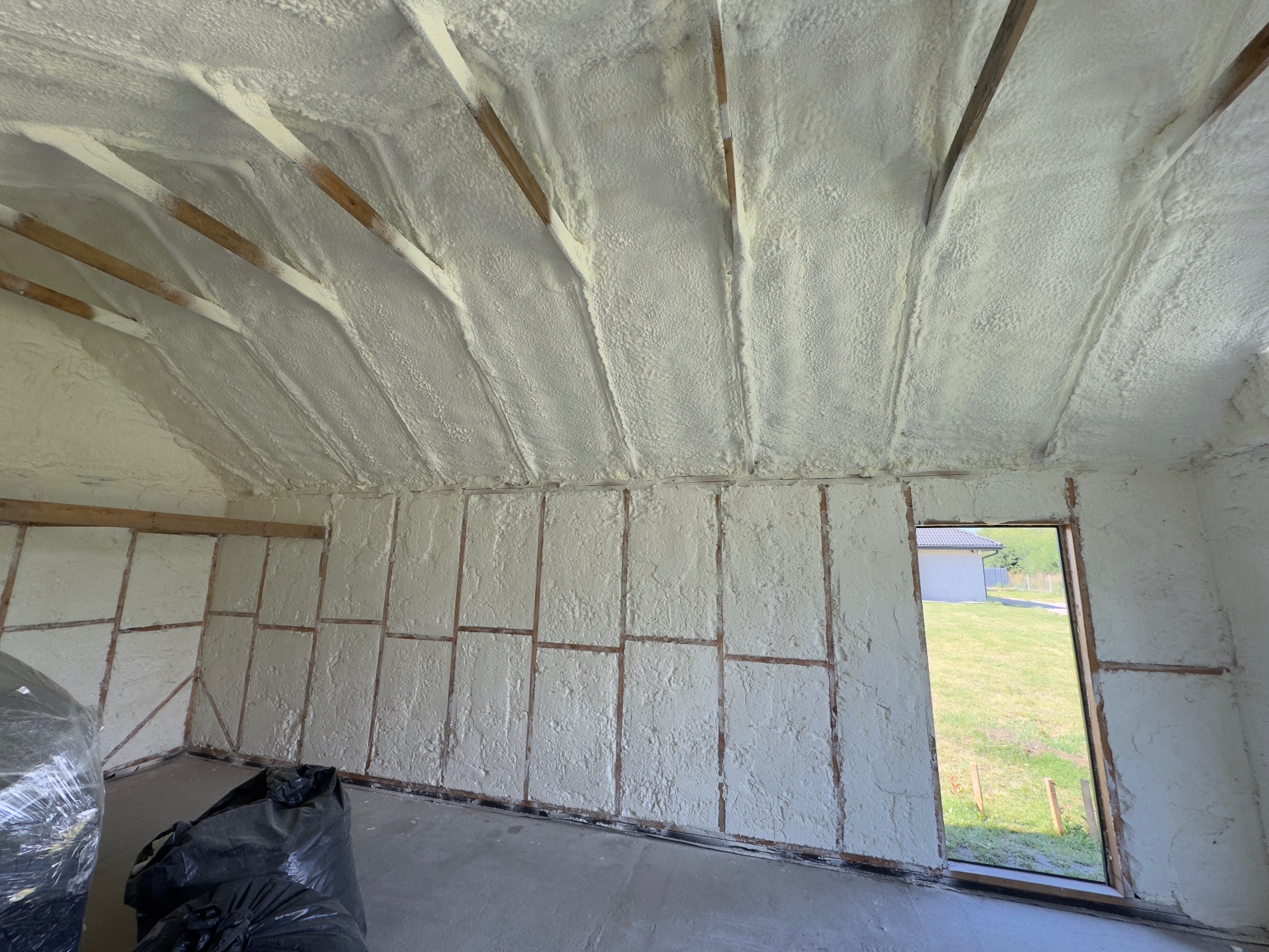 Poddasze po natrysku izolacji. Widoczne drewniane elementy konstrukcji i równomiernie nałożona warstwa izolacji. W tle okno z widokiem na zewnątrz.