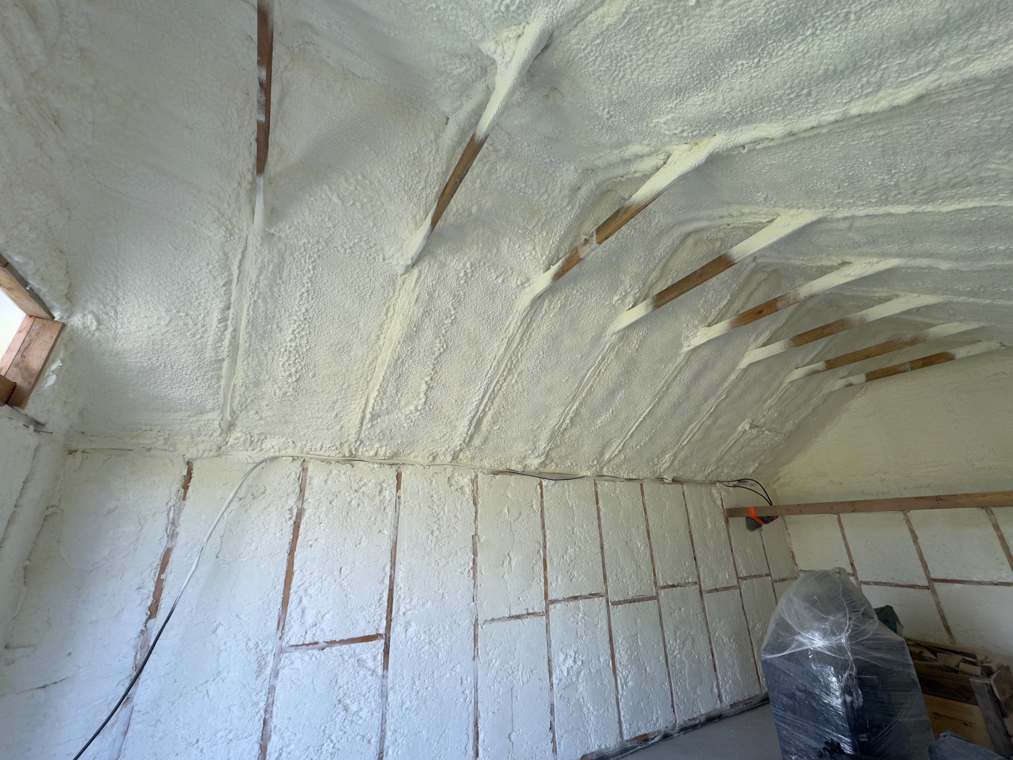 Poddasze po natrysku izolacji. Widoczne drewniane elementy konstrukcyjne i równomierna warstwa białej piany izolacyjnej na ścianach i suficie. Izolacja wykonana natryskowo.