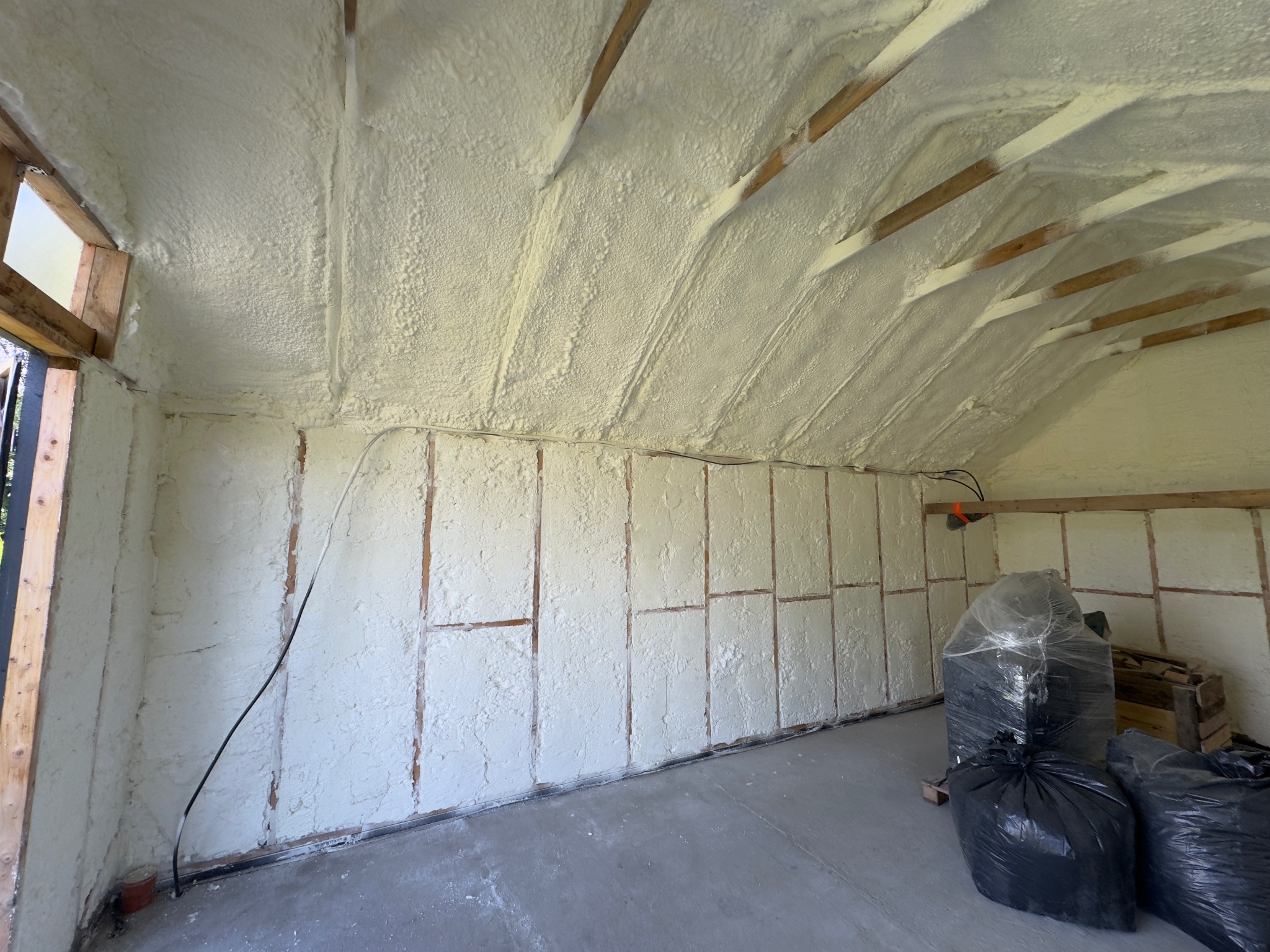 Poddasze po natrysku izolacji. Widoczne drewniane elementy konstrukcyjne i równomiernie nałożona warstwa izolacji. W rogu worki z odpadami budowlanymi.