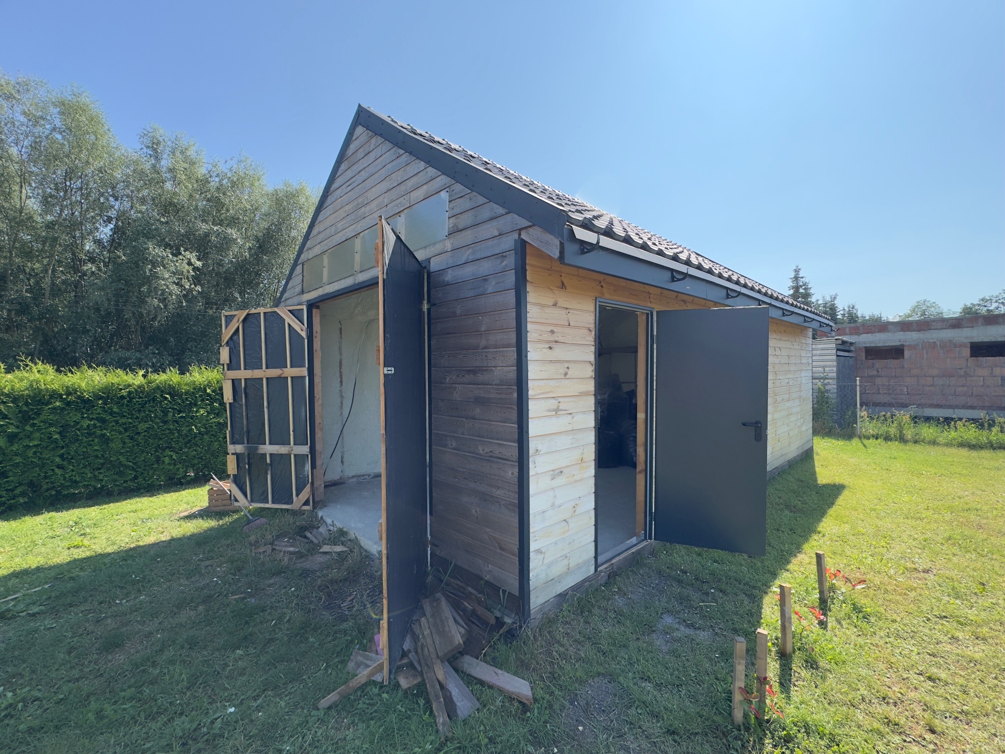 Otwarty budynek gospodarczy z widoczną izolacją natryskową, drewniana elewacja, czarne drzwi, trawnik i błękitne niebo w tle. Widoczny etap prac.