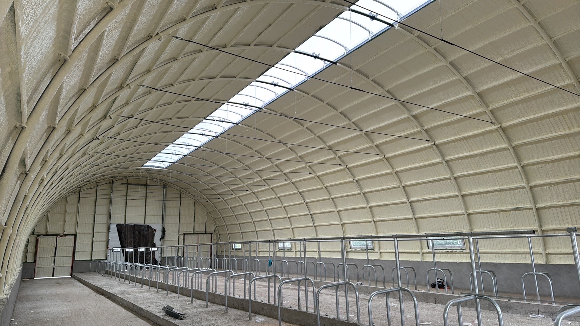 Wnętrze ocieplonego pianką PUR budynku o łukowej konstrukcji dachu z świetlikami. Widoczne metalowe elementy konstrukcyjne i ściany pokryte jasną pianą.