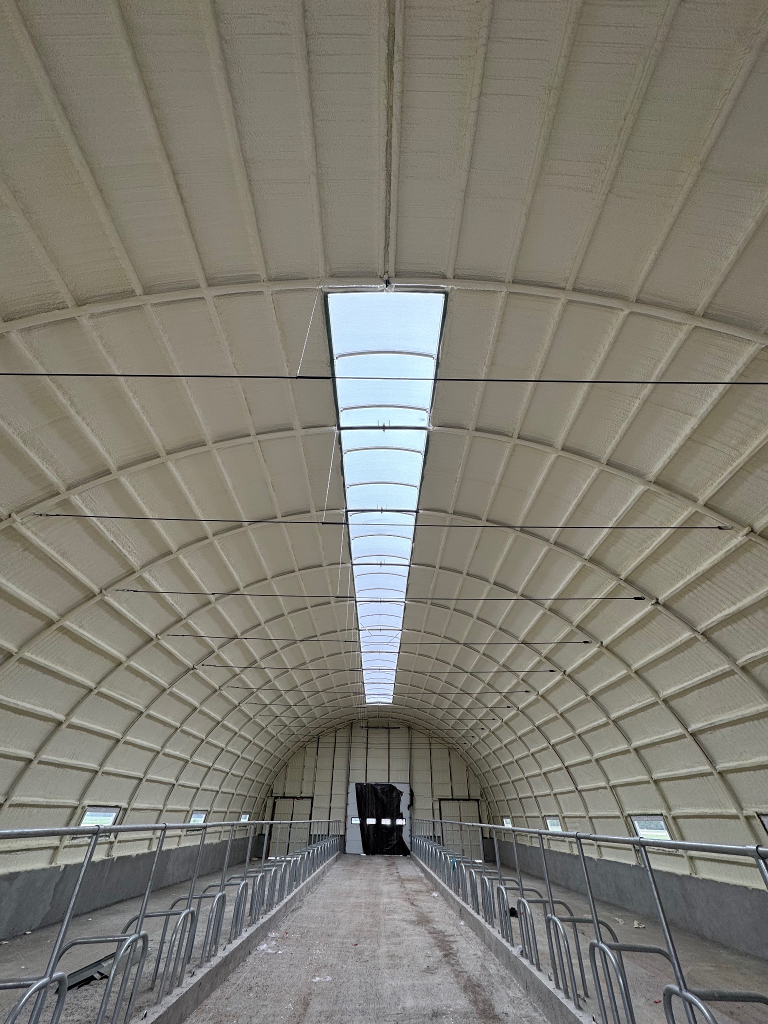 Wnętrze hali z ocieplonym pianką PUR dachem w kształcie łuku, widok z perspektywy posadzki, z oknami w górnej części ścian i świetlikiem w dachu. Metalowe barierki po bokach.