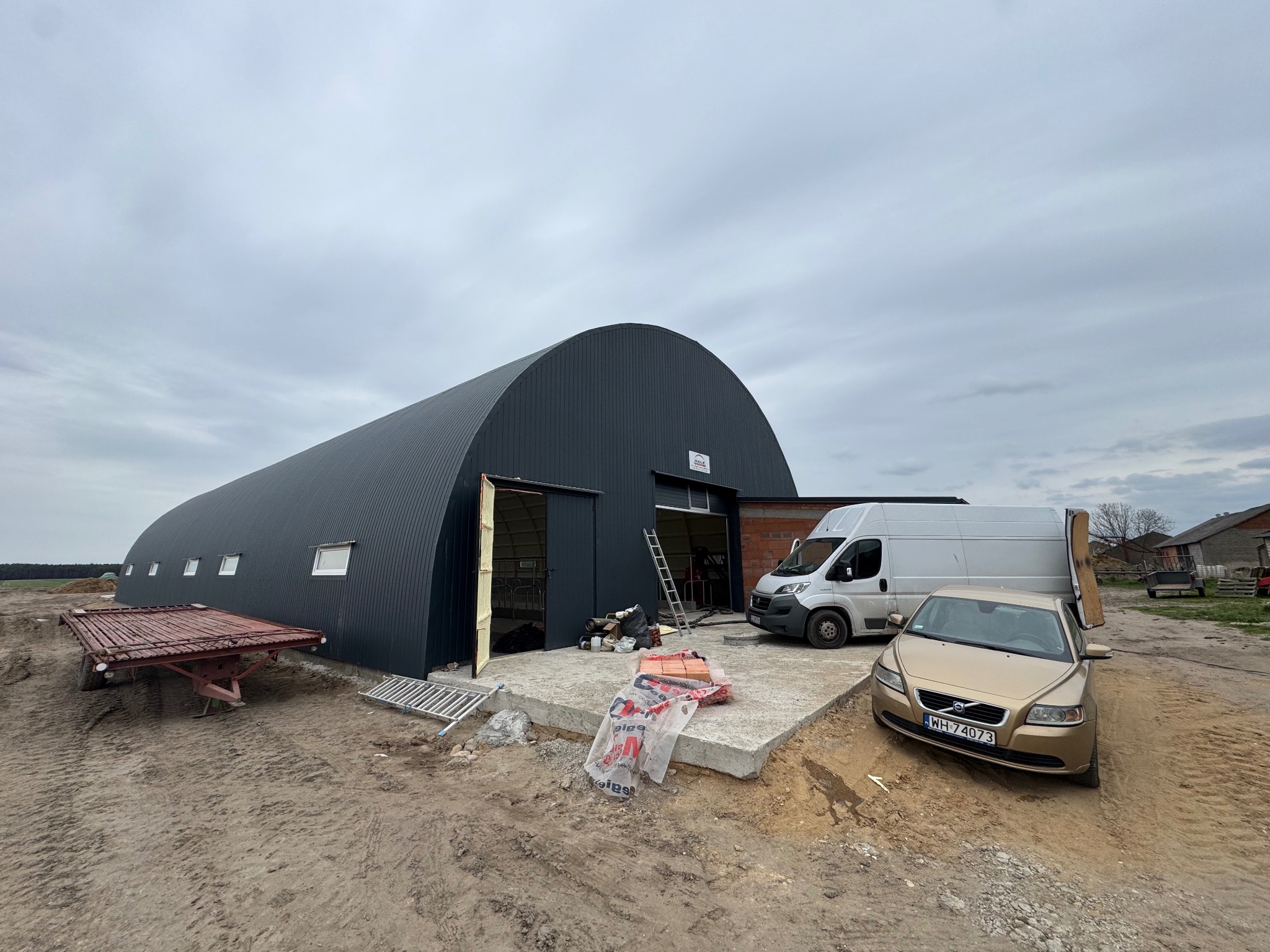 Ocieplony, łukowy budynek gospodarczy z blachy trapezowej w kolorze grafitowym. Na pierwszym planie samochód osobowy i dostawczy. Widoczne materiały budowlane i drabina.