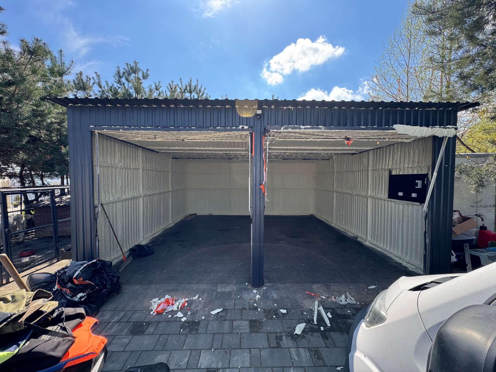Ocieplony garaż blaszany z otwartymi wrotami, widoczne ślady pianki na podłożu. W tle zieleń drzew i błękitne niebo. Ujęcie z zewnątrz, z bliska.