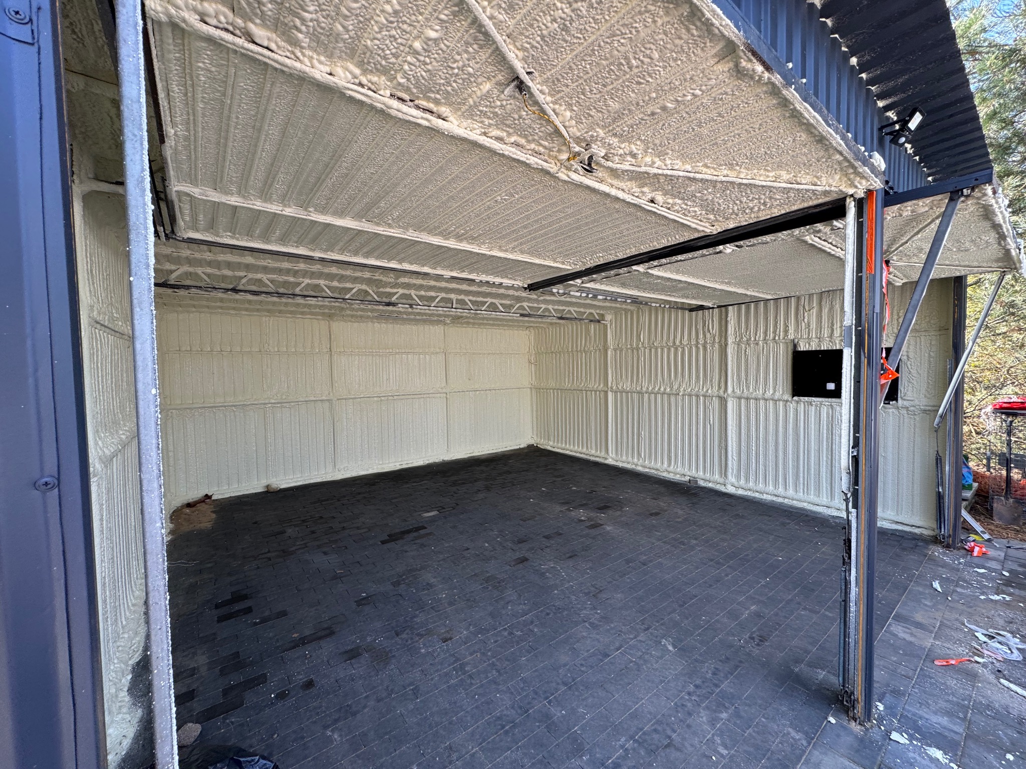 Wnętrze garażu po natrysku izolacji. Biała pianka pokrywa ściany i sufit, tworząc jednolitą warstwę. Widoczna czarna podłoga z płytek i metalowa konstrukcja dachu.