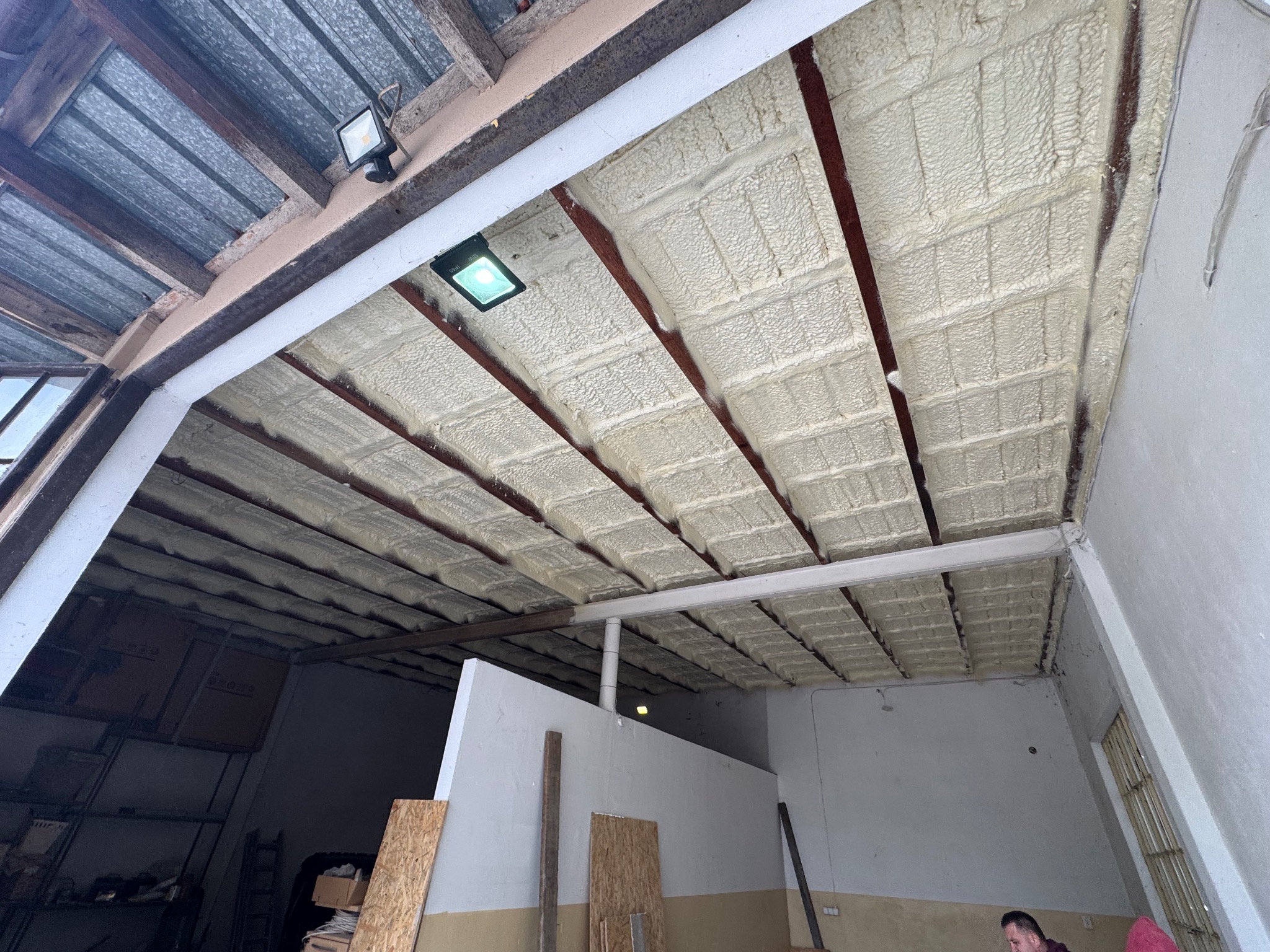 Ocieplenie poddasza pianką PUR, widoczne równomierne pokrycie między krokwiami. Oświetlenie LED w tle. Częściowo widoczne płyty OSB i fragment sylwetki osoby.