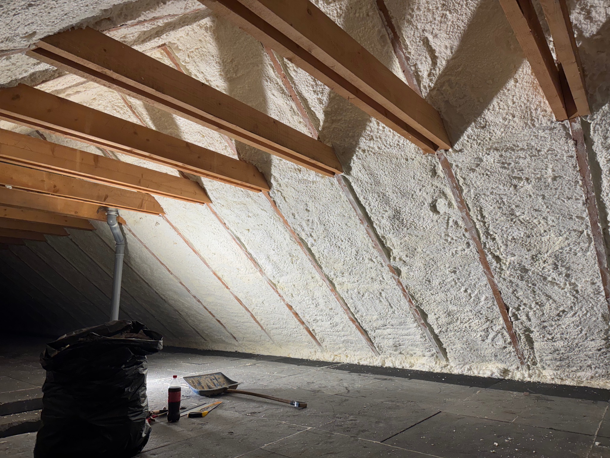 Poddasze po natrysku izolacji: drewniane belki konstrukcyjne otulone grubą warstwą jasnej piany. Na podłodze narzędzia i worek, co sugeruje etap końcowy prac.