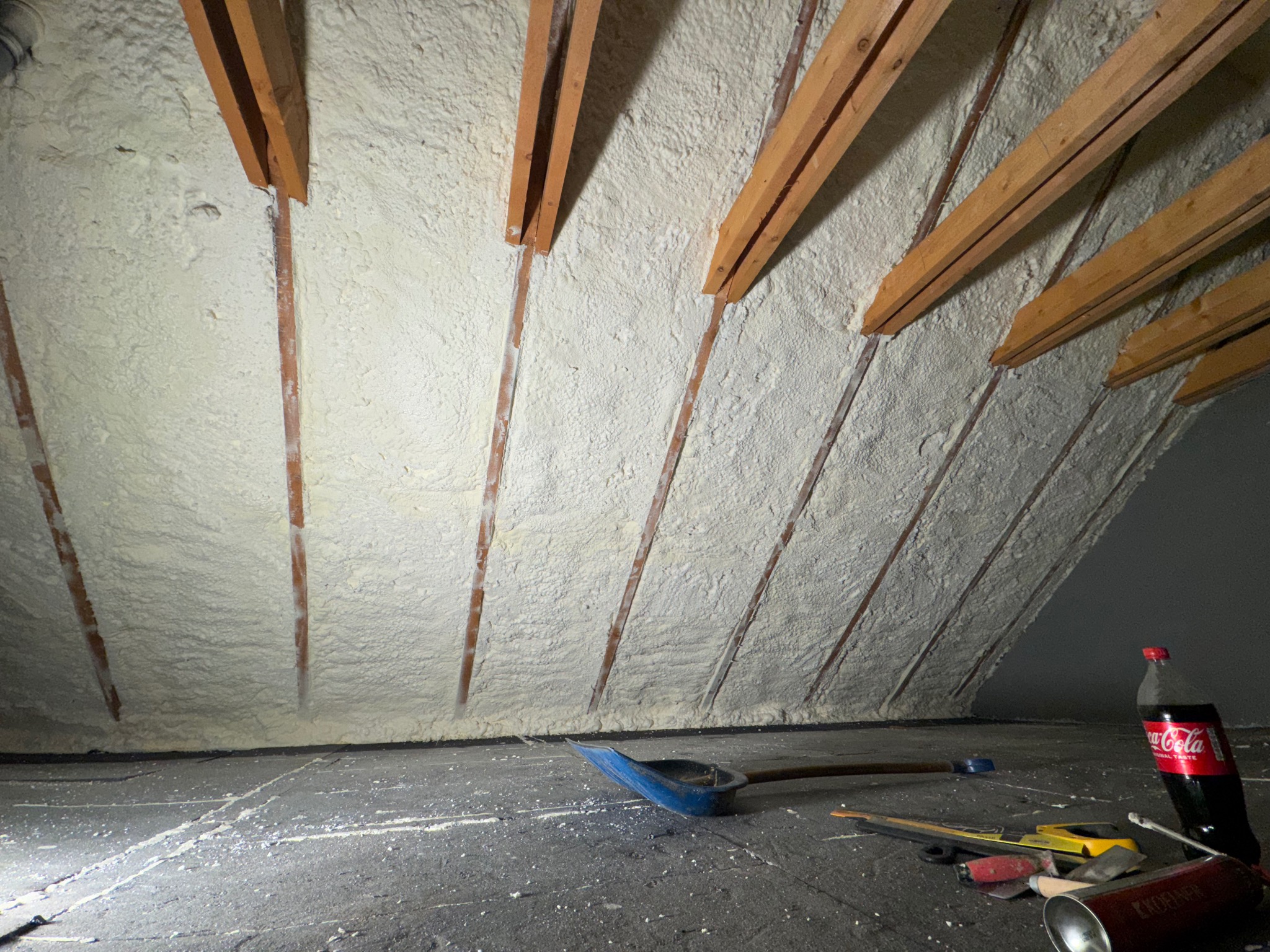 Poddasze po natrysku izolacji piankowej, widoczne drewniane krokwie i narzędzia pracy na podłodze. Izolacja o jednolitej strukturze pokrywa całą powierzchnię.