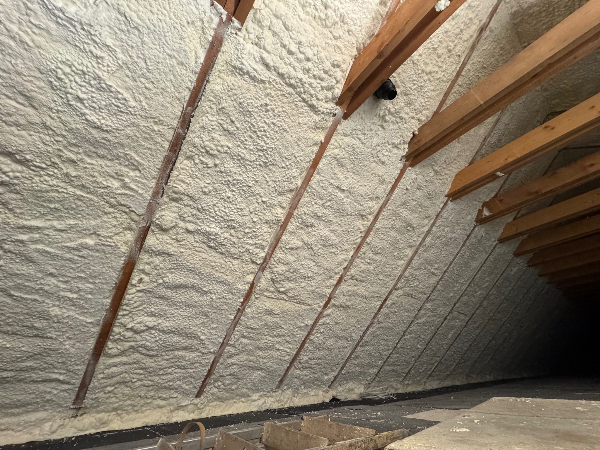 Poddasze po natrysku izolacji. Widoczne drewniane krokwie i równomierna warstwa piany izolacyjnej, szczelnie wypełniająca przestrzeń. Izolacja w kolorze kremowym.