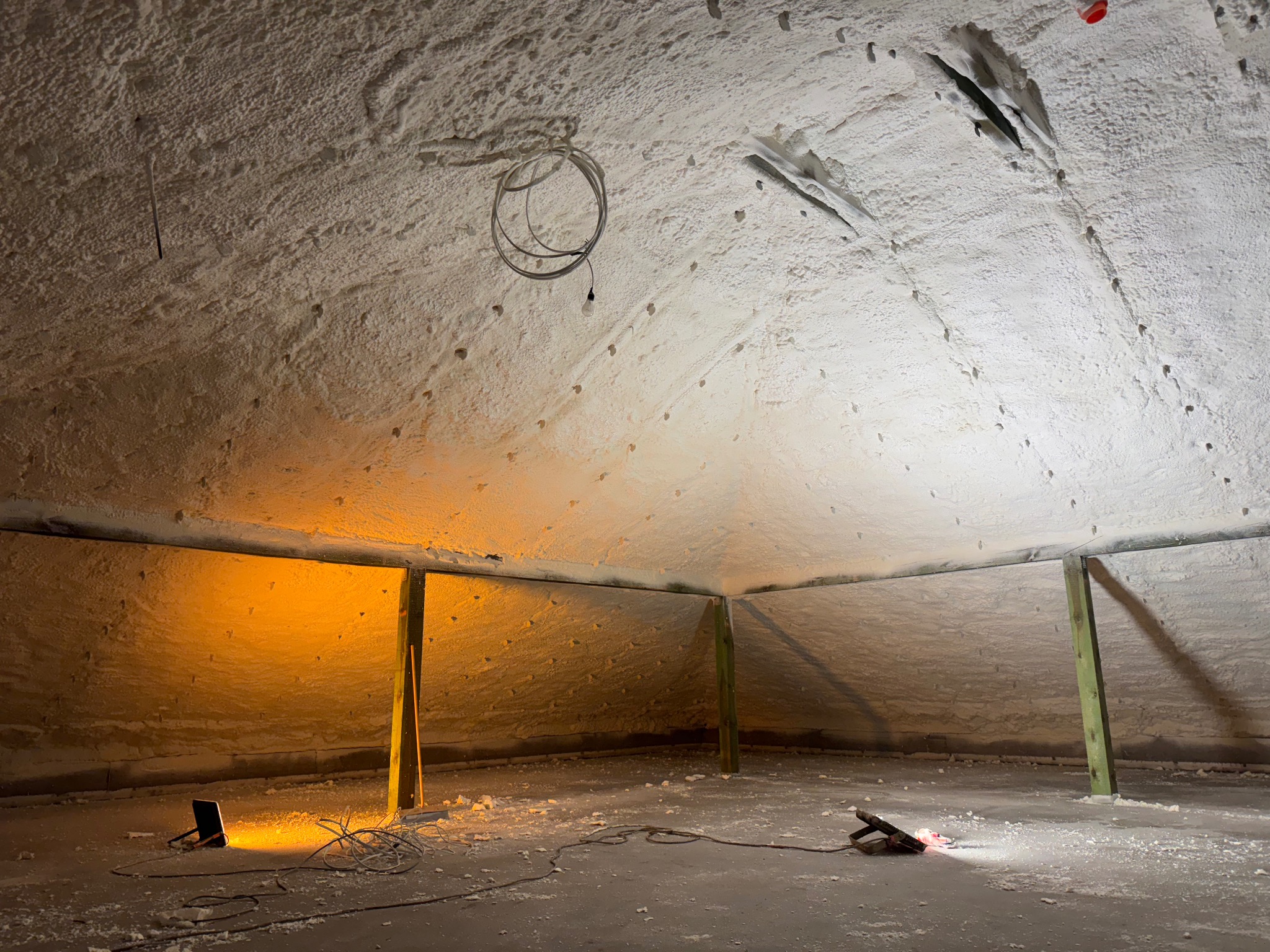 Poddasze po natrysku izolacji. Widoczne drewniane elementy konstrukcyjne, oświetlenie robocze i przewody. Biała, porowata powierzchnia izolacji pokrywa ściany i sufit.