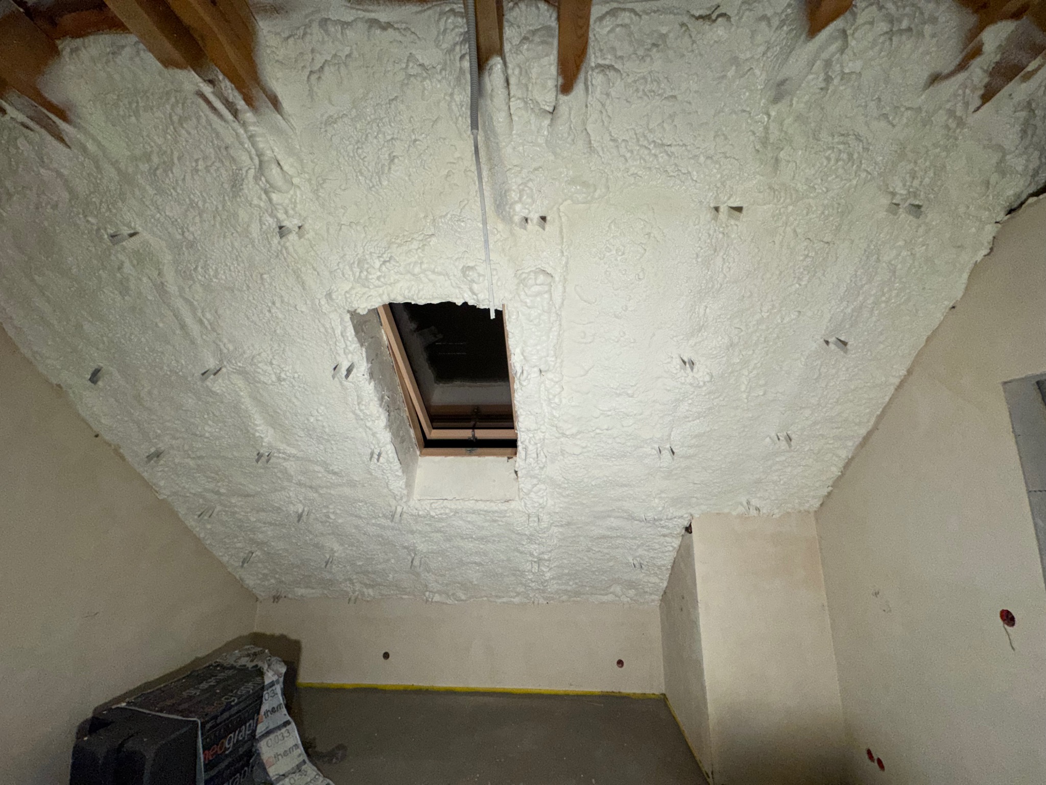 Poddasze po natrysku izolacji pianką PUR, widoczne drewniane elementy konstrukcyjne i okno dachowe. Biała, porowata struktura pianki pokrywa skosy i sufit.