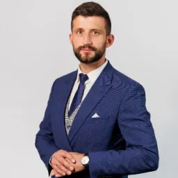 Portret mężczyzny w granatowym garniturze z kamizelką w kratę, zegarkiem na ręku i obrączką, na jasnym tle.