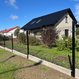 FENSIS- ogrodzenia systemowe Rafał Galas - Czarny panelowy płot na betonowej podmurówce, oddzielający posesję z nowoczesnym domem z czarną dachówką od trawnika, w tle inne domy i błękitne niebo.