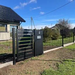 Nowoczesna czarna brama wjazdowa z poziomymi panelami, zintegrowana z ogrodzeniem z siatki na betonowej podmurówce przed domem z ciemną dachówką w słoneczny dzień.