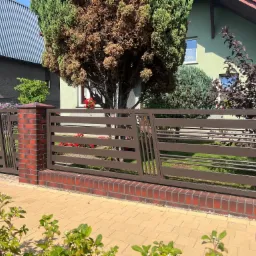 Nowoczesne, poziome przęsła z brązowego metalu tworzą ogrodzenie z czerwonej cegły, z roślinnością w tle i żółtą kostką brukową na pierwszym planie.