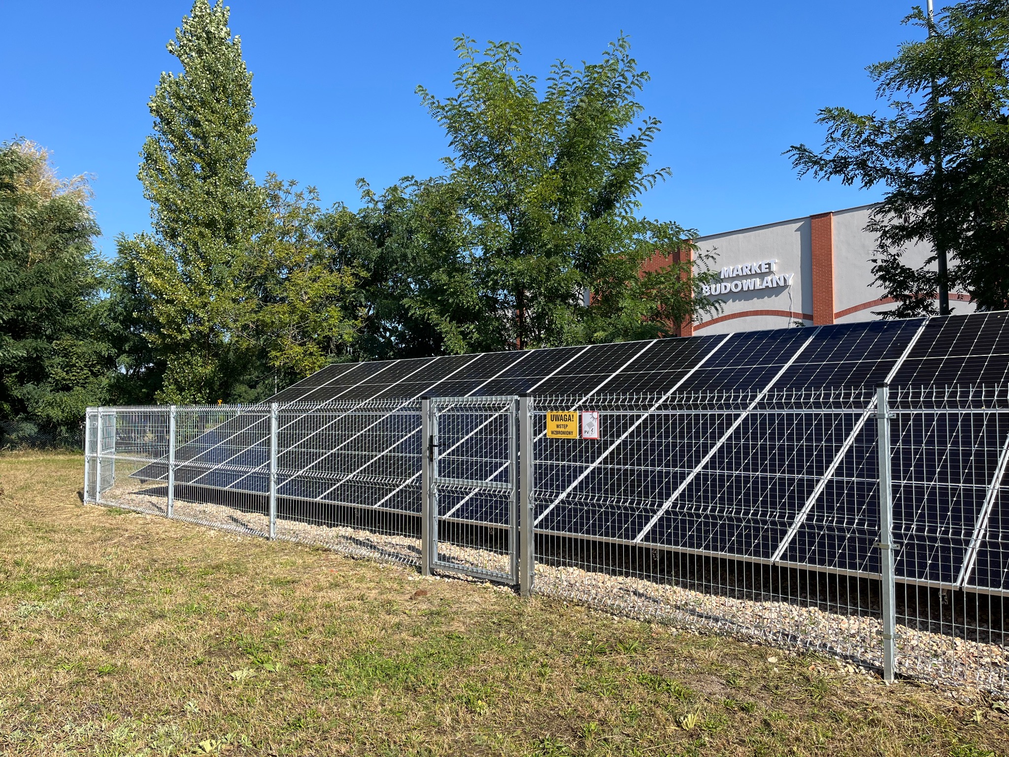 Instalacja paneli słonecznych zabezpieczona metalową siatką z bramką, teren zielony, w tle budynek marketu budowlanego pod błękitnym niebem.