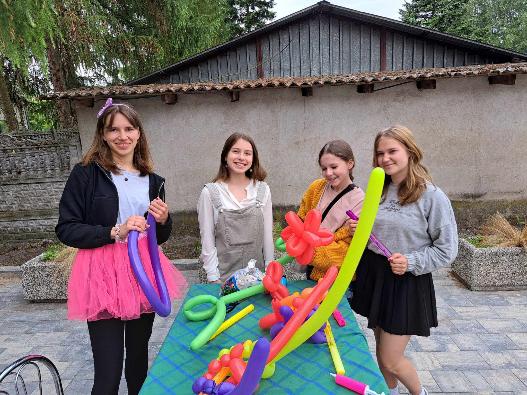 Cztery uśmiechnięte dziewczyny prezentują kolorowe balony modelowane w różne kształty na zielonym stole, na zewnątrz, przy budynku.