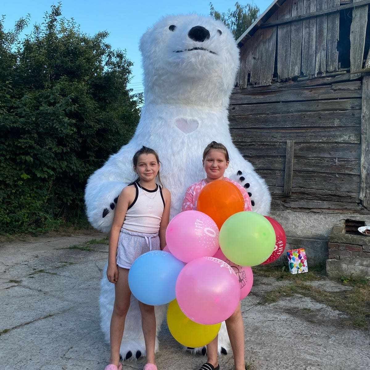 Dwie dziewczynki trzymające balony pozują z animatorem przebranym za białego niedźwiedzia na tle drewnianego budynku.