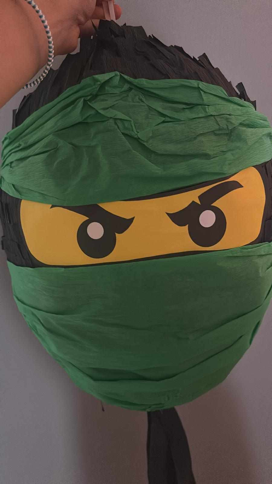 Piñata w kształcie głowy zielonego ninja, wykonana z krepy i kartonu, z charakterystycznymi żółtymi oczami.