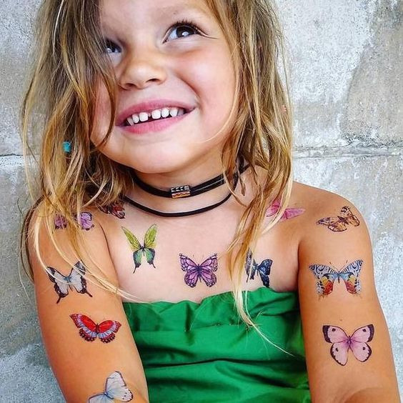 Uśmiechnięta dziewczynka z tymczasowymi tatuażami motyli na ramionach i dekolcie, ubrana w zieloną sukienkę i czarny choker.