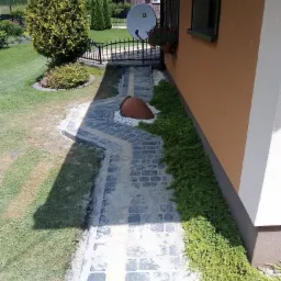 Ścieżka z kostki brukowej prowadząca wzdłuż ściany domu, z dekoracyjnym dzbanem na białych kamieniach i niskimi roślinami przy ścianie.