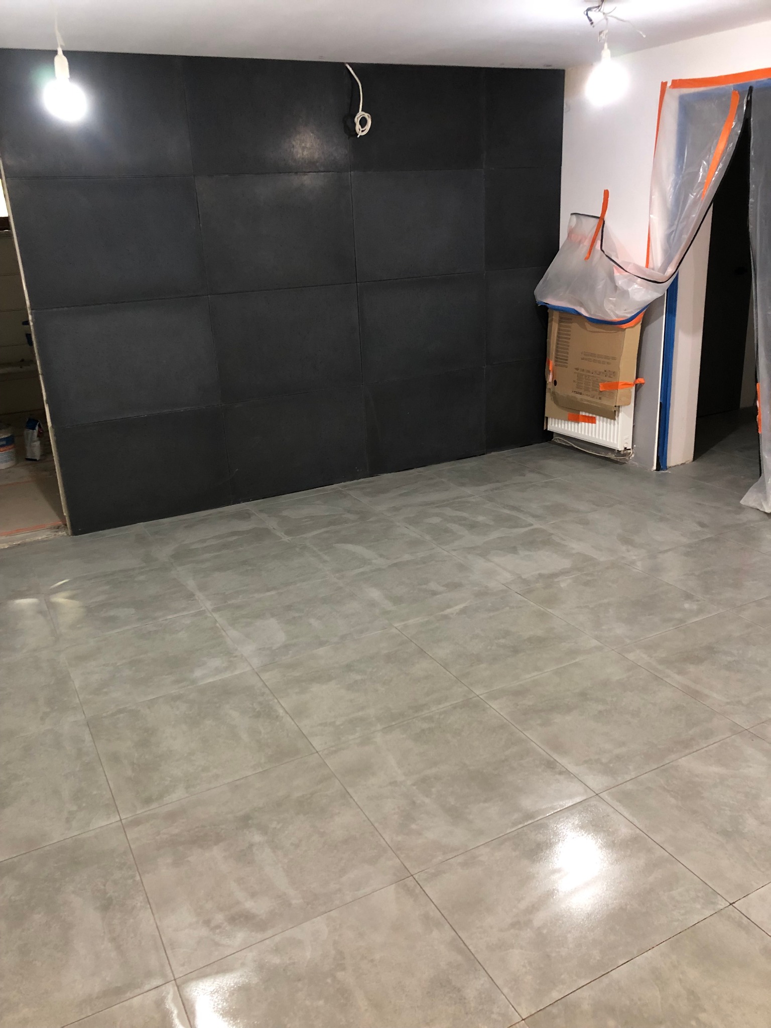 Łazienka w trakcie remontu: ułożone szare płytki podłogowe, ściana wyłożona ciemnymi panelami, zabezpieczone folią drzwi.