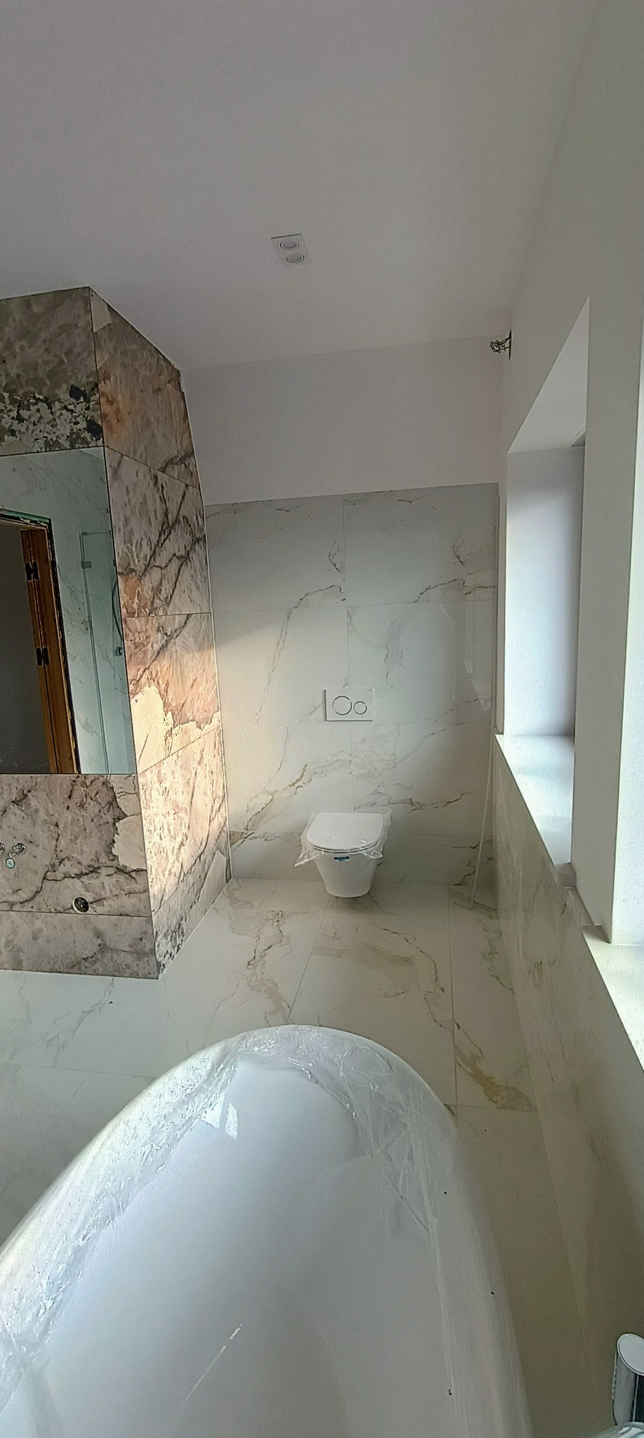 Nowoczesna łazienka z marmurowymi płytkami i podwieszaną toaletą, wanna zabezpieczona folią ochronną.