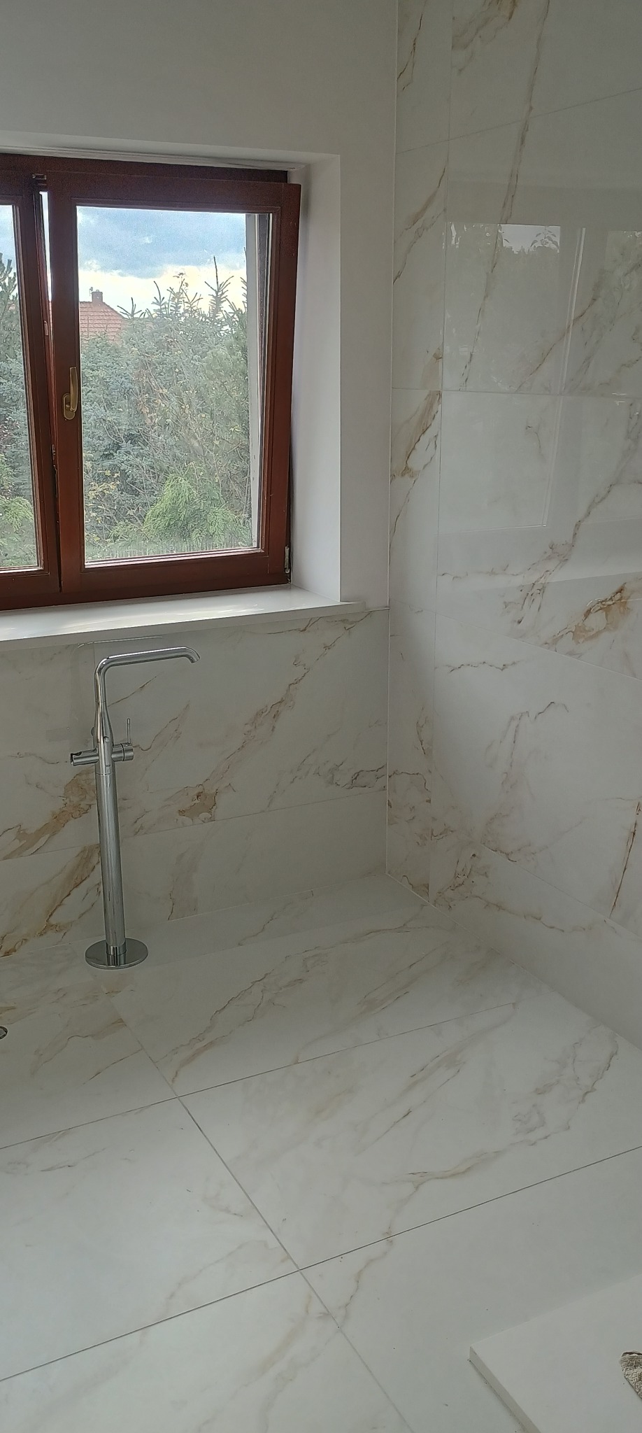 Wykończona łazienka z białymi płytkami imitującymi marmur, brązowe okno z widokiem na zieleń i chromowana bateria podłogowa.