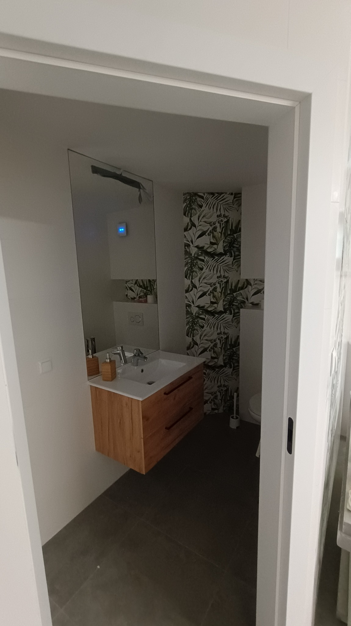 Nowoczesna łazienka z drewnianą szafką pod umywalką, wzorzystą tapetą w liście i dużym lustrem, widok zza framugi drzwi.