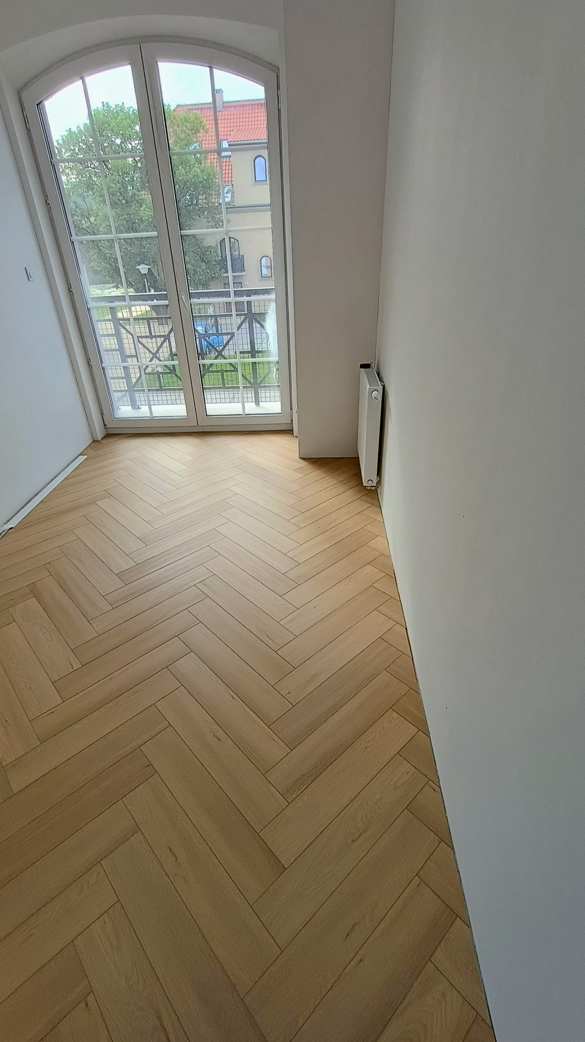 Nowa podłoga ułożona w jodełkę w jasnym wnętrzu we Wrocławiu, widok na okno balkonowe i grzejnik.