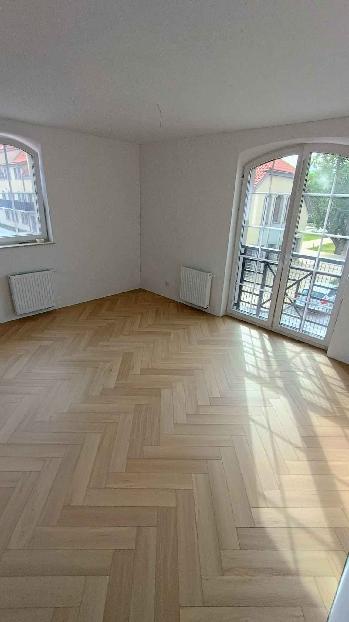 Jasny parkiet ułożony we wzór jodełki francuskiej w pustym, jasnym pokoju z dwoma oknami łukowymi i grzejnikami pod oknami.
