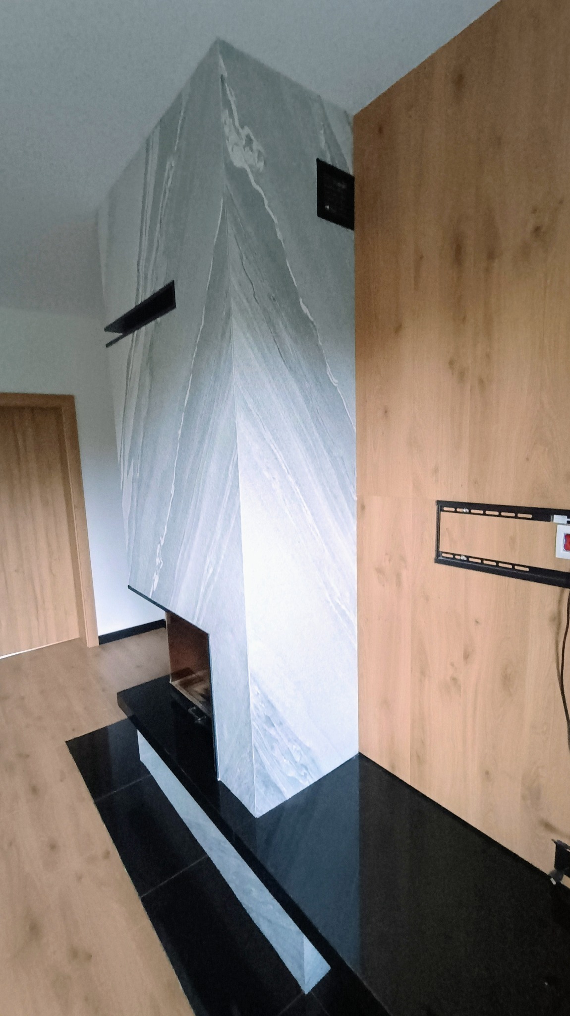 Nowoczesny kominek obudowany płytami z szarego marmuru z białymi żyłami, z czarnym, błyszczącym blatem i drewnianą ścianą w tle, z zamontowanym uchwytem do telewizora.