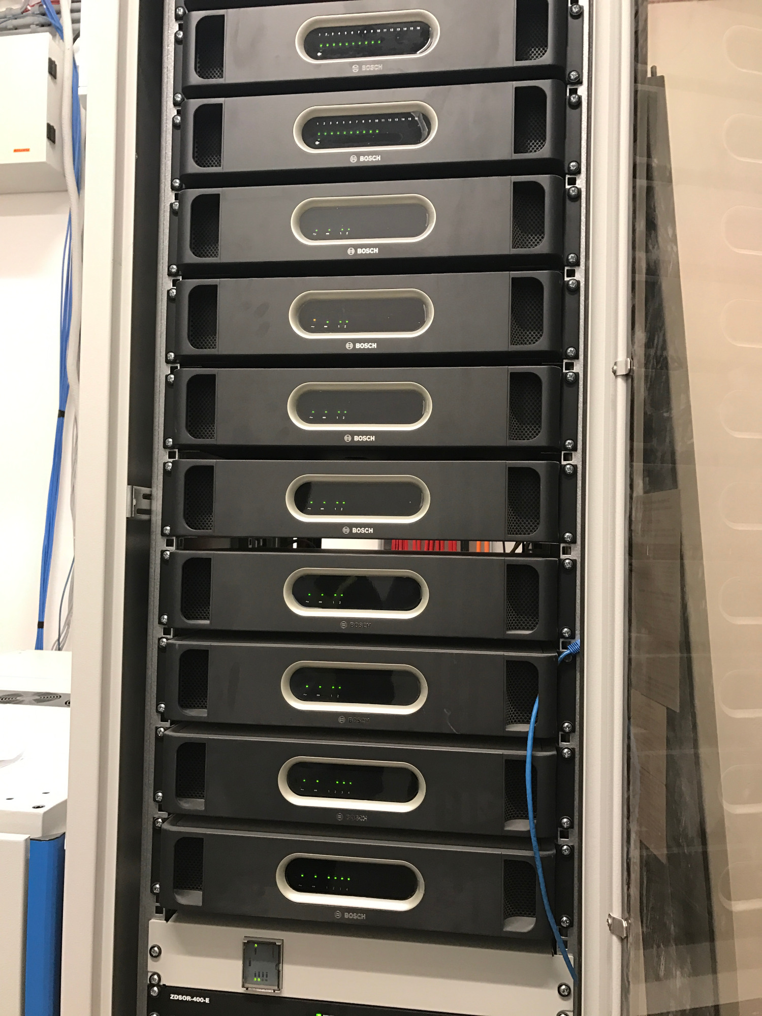 Rząd czarnych urządzeń Bosch w szafie rack, z widocznymi diodami LED i niebieskim kablem Ethernet, zainstalowane w pomieszczeniu z białymi ścianami.