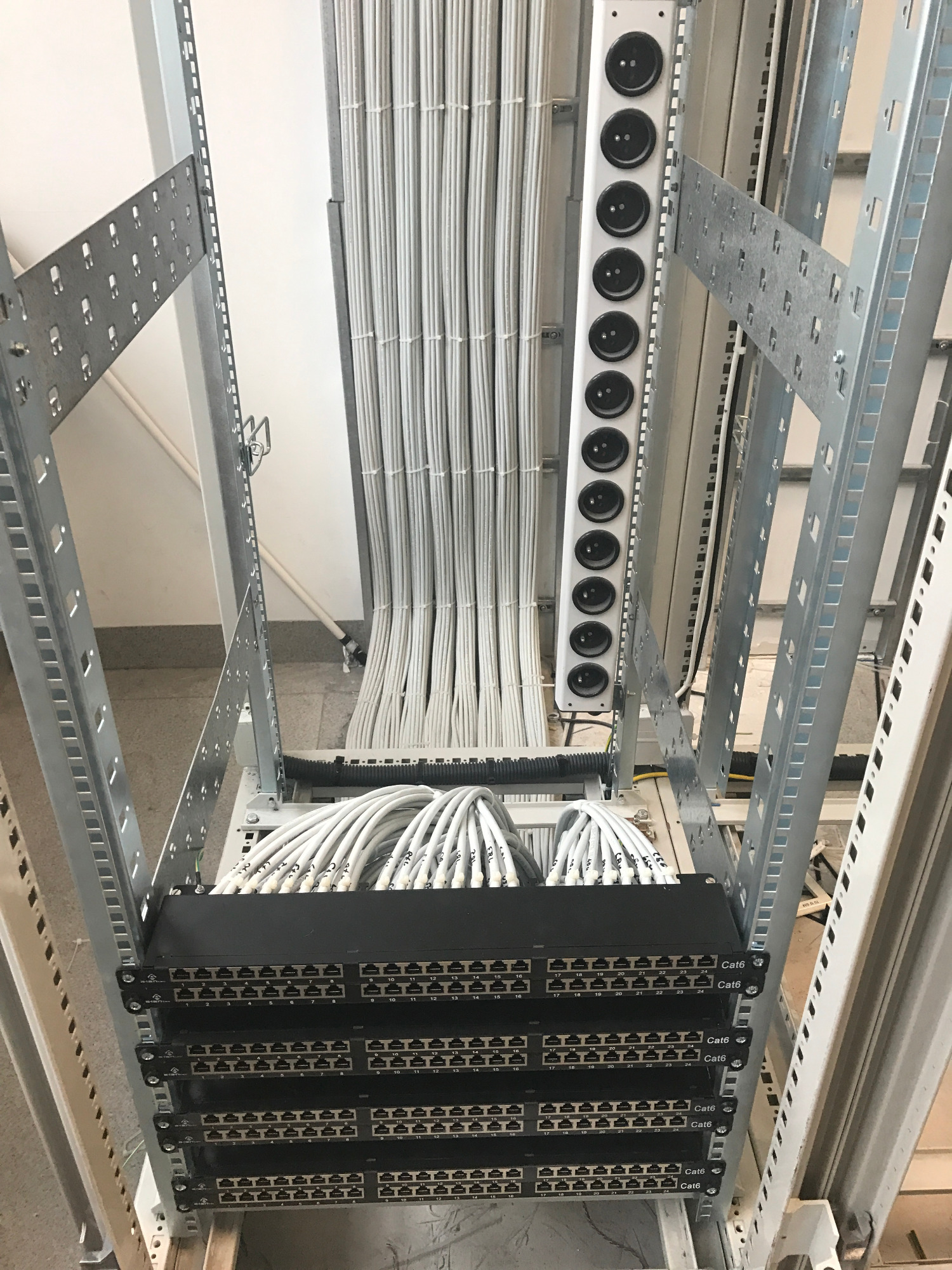 Rządowe kable sieciowe Cat6 uporządkowane w szafie rack, z widocznymi panelami krosowniczymi i listwą zasilającą z czarnymi gniazdami, całość w surowym, technicznym otoczeniu serwerowni.